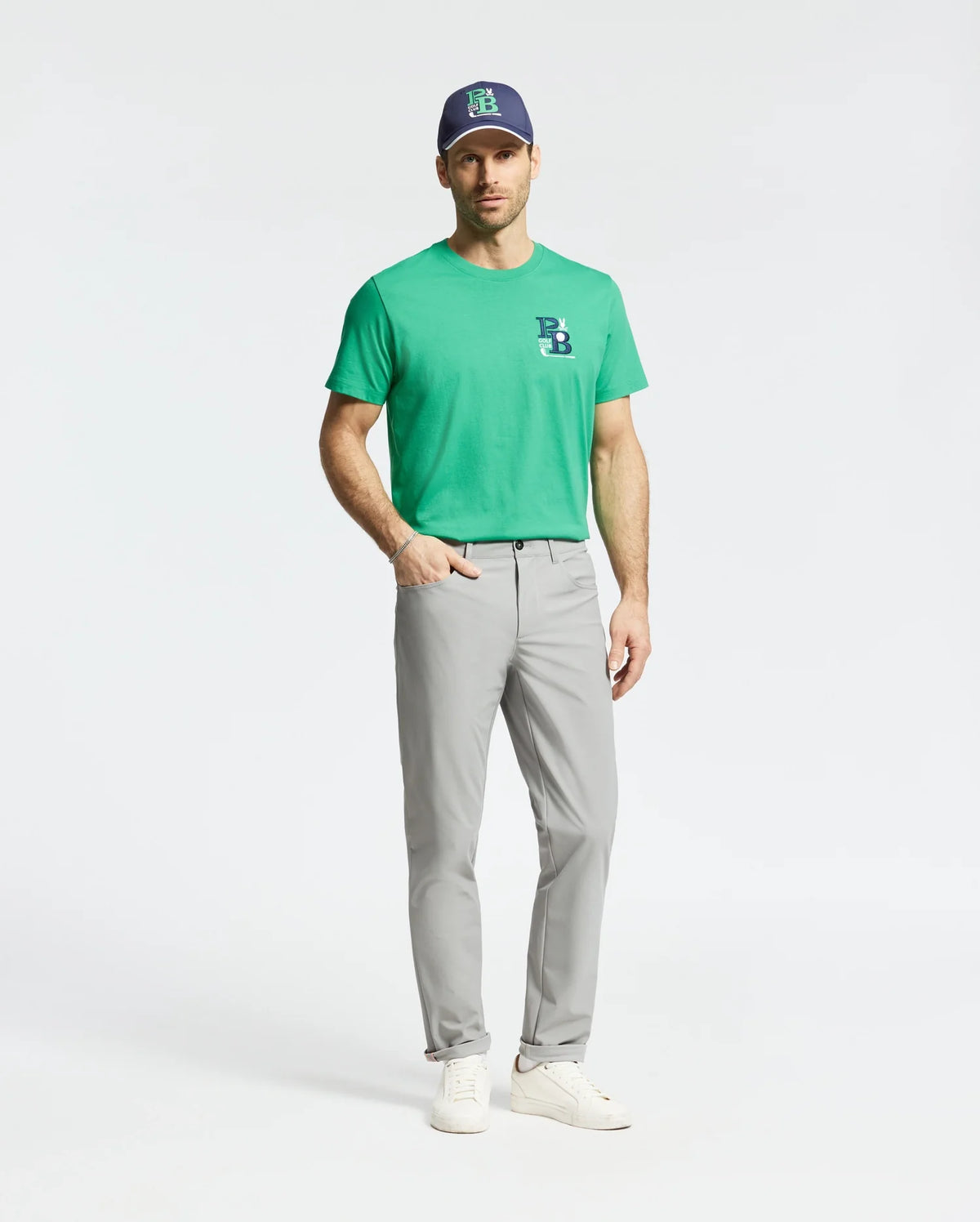 A man stands against a plain background wearing Psycho Bunny's MENS DENIS GRAPHIC TEE (MT0200015) in golf-inspired green, crafted from soft Pima cotton with an embroidered logo, light gray pants, a navy cap featuring a green logo, and white sneakers.
