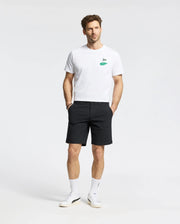 A man in the Psycho Bunny MENS DALE GRAPHIC TEE (MT0200016), a white Pima cotton T-shirt with a green embroidered logo, pairs it with black shorts. He wears white sneakers and socks, posing against a plain background with understated yet refined casual elegance.