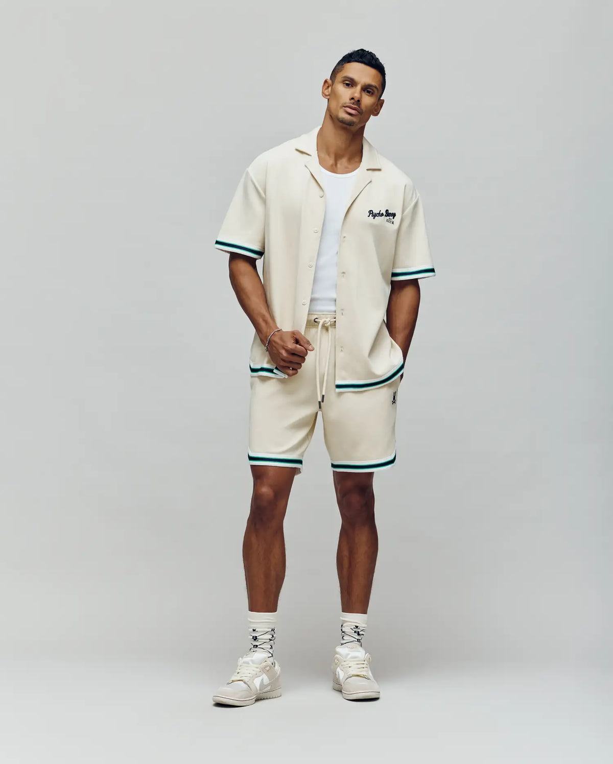 A man stands against a plain background wearing the Psycho Bunny MENS BASIL TAPED CAMP COLLAR SHIRT (MT0500004), matching shorts with green trim, a white tank, white socks, and cream sneakers. One hand is in his pocket.