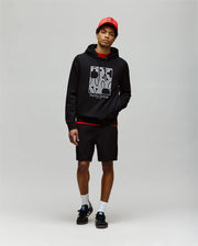 A young man stands against a plain background wearing the Psycho Bunny MENS GLENN HOODIE - MT0500008, featuring an embroidered Bunny graphic, paired with black shorts, black sneakers, white socks, and a red baseball cap. His left hand is in his pocket.