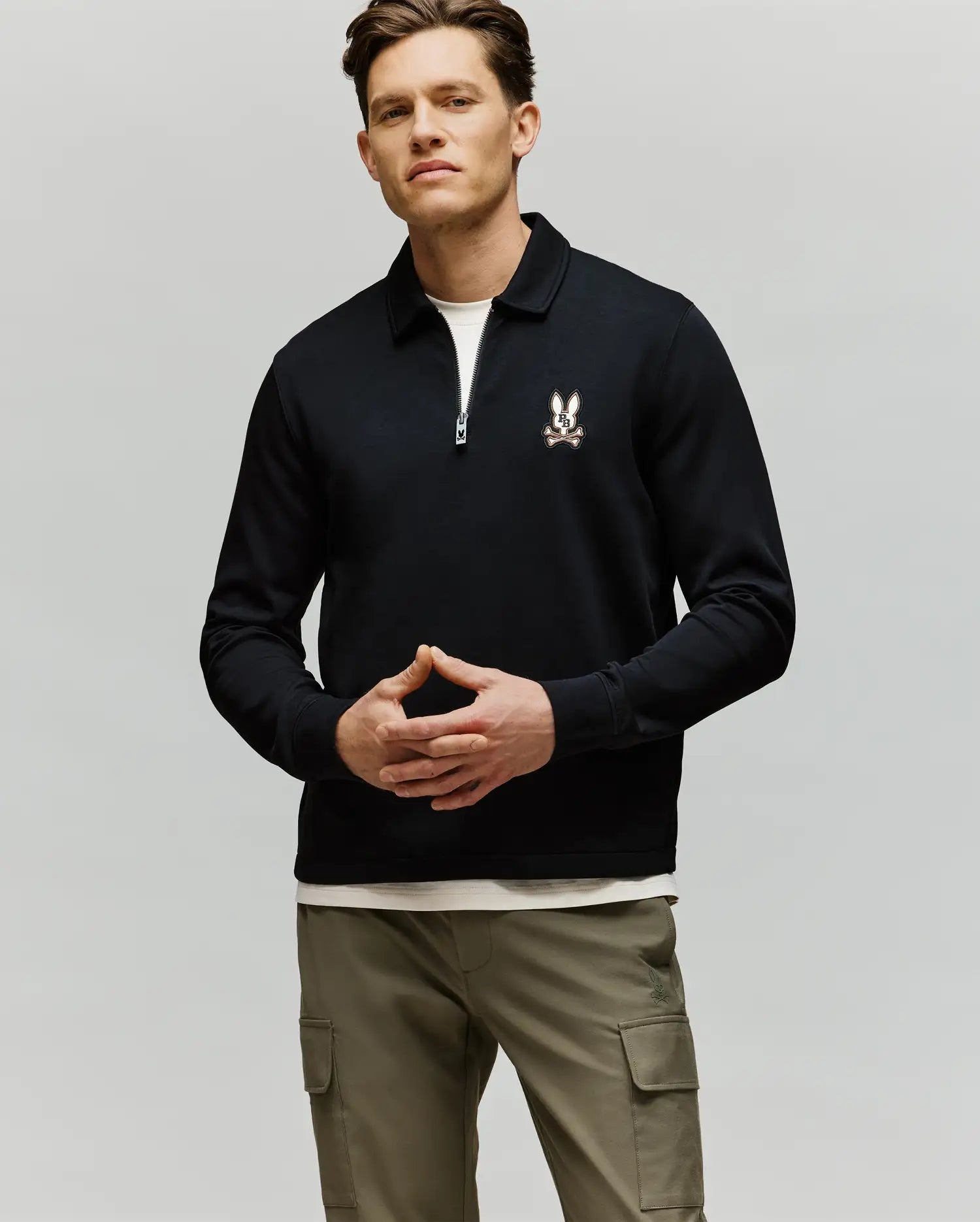 A man stands against a plain background wearing the Psycho Bunny MENS DANIEL QUARTER ZIP SWEATSHIRT - MT0500023 with a small logo on the left chest, light cargo pants, and a white shirt peeking from under the sweatshirt.