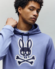 Wearing the Psycho Bunny MENS RYDER BUNNY HOODIE (MT0500029) in light purple, featuring a bold skull-and-crossbones with bunny ears graphic, a confident person poses with one hand near their neck—perfect for stylish lounging.