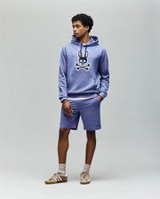 A person stands against a plain background wearing the Psycho Bunny MENS RYDER BUNNY HOODIE (MT0500029) in lavender with an embroidered Bunny patch and cartoon crossbones, matching loungewear shorts, beige socks, and tan sneakers.