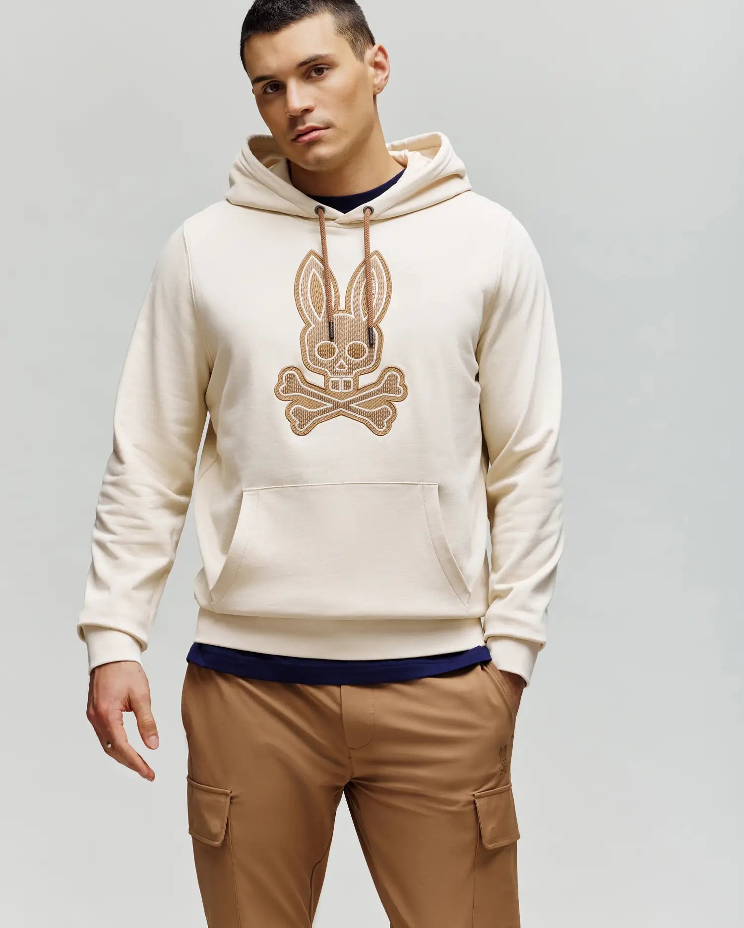 A man wears the Psycho Bunny MENS RYDER BUNNY HOODIE (MT0500029) in cream, featuring an embroidered Bunny patch and cartoon crossbones graphic, styled with tan cargo pants against a light gray background—ideal for cozy loungewear.