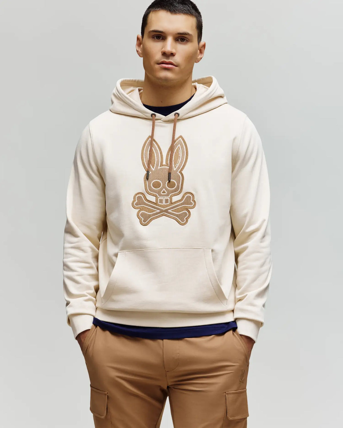 A man with short dark hair wears the Psycho Bunny MENS RYDER BUNNY HOODIE (MT0500029) in cream, featuring a large embroidered Bunny patch with crossbones on the front, along with tan loungewear pants, against a light gray background.