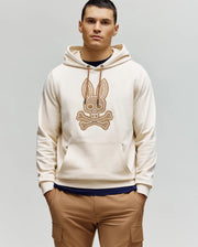 A man with short dark hair wears the Psycho Bunny MENS RYDER BUNNY HOODIE (MT0500029) in cream, featuring a large embroidered Bunny patch with crossbones on the front, along with tan loungewear pants, against a light gray background.