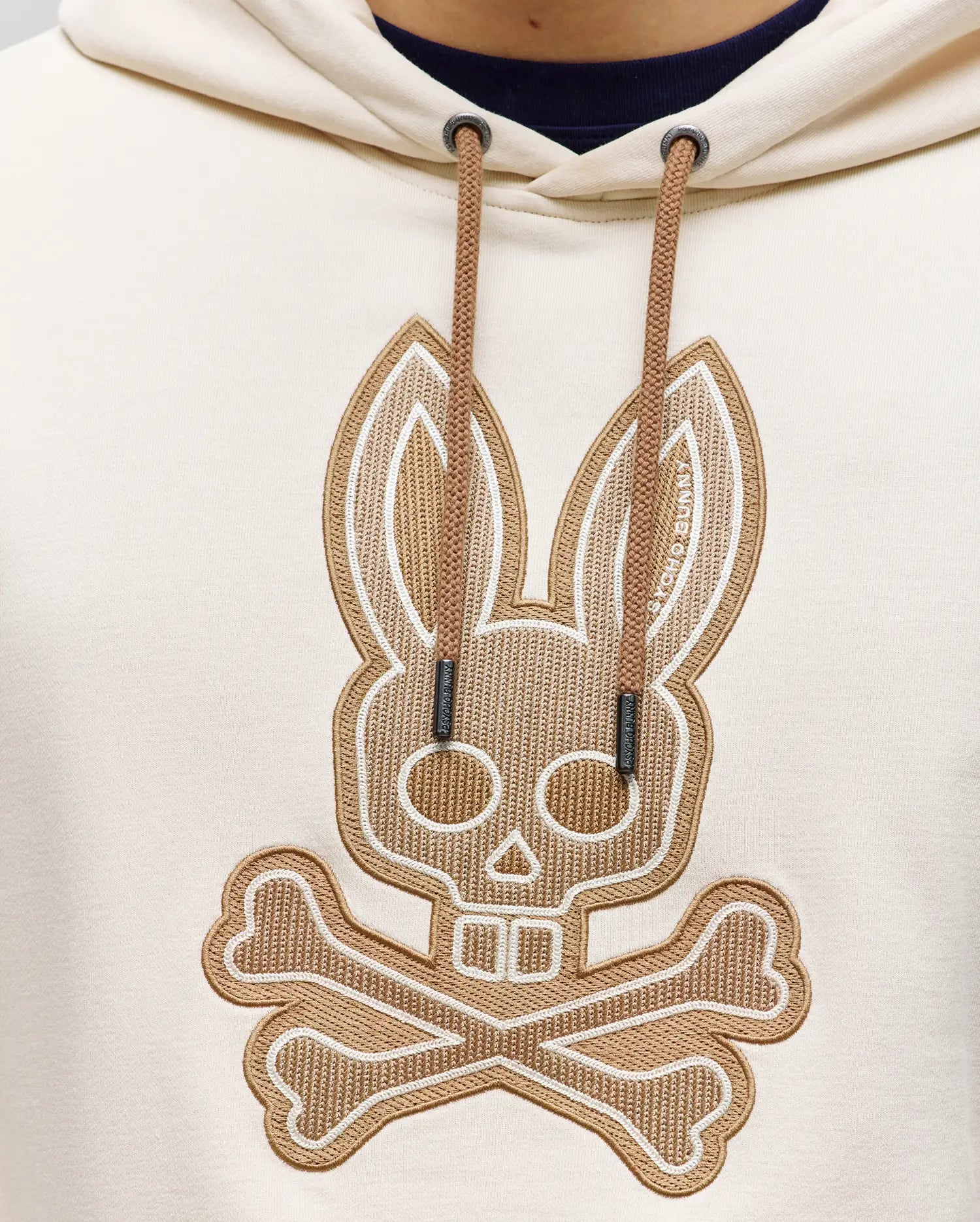 Close-up of the cream-colored Psycho Bunny MENS RYDER BUNNY HOODIE (MT0500029), featuring an embroidered bunny patch—skull with rabbit ears over crossed bones—for a playful spin on the classic skull and crossbones. Perfect standout loungewear.