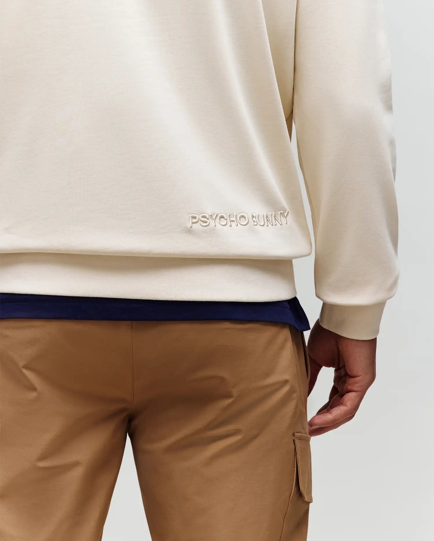 A person is seen from behind wearing the Psycho Bunny MENS RYDER BUNNY HOODIE (MT0500029) in cream, featuring an embroidered Bunny patch near the hem, styled with tan cargo pants for a relaxed loungewear look.