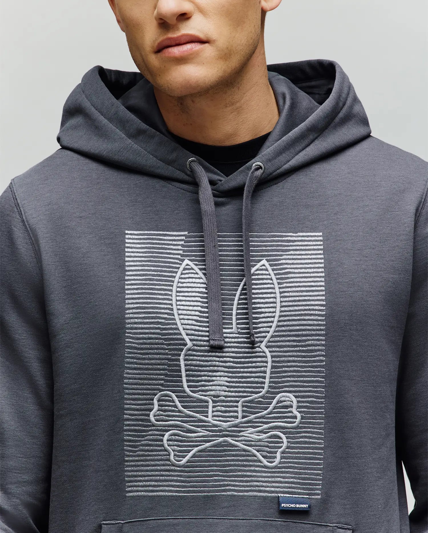 A person wears the Psycho Bunny MENS GRANT HOODIE - MT0500032, a gray premium cotton blend featuring a playful white zig-zag embroidered bunny head above crossed bones. Their face is partially visible.
