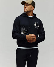 A man in the Psycho Bunny MENS AUGUSTE HOODIE (MT0500043) and matching joggers—both with embroidered bunny and "PB" logos—wears a green-brown cap, looking to the side against a plain background.
