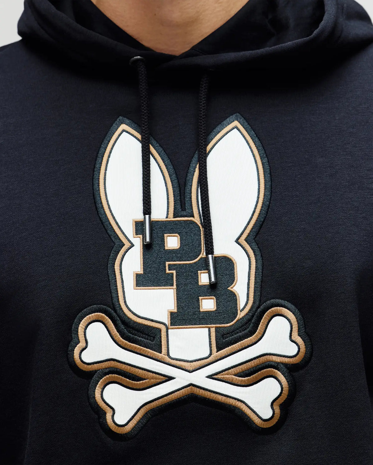 A close-up of the Psycho Bunny MENS WADE HOODIE - MT0500112 in black, showcasing an embroidered Bunny patch with ears, PB initials, and crossed bones, crafted from a premium cotton blend.