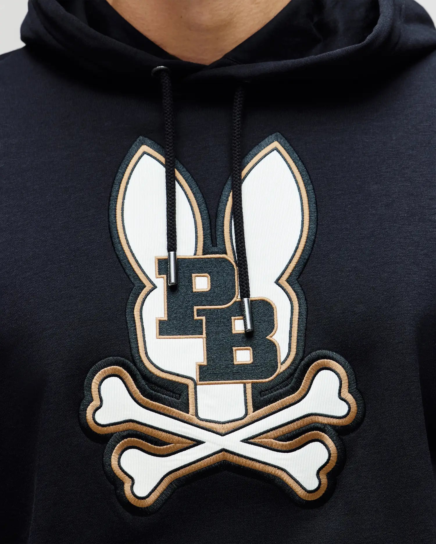 A close-up of the Psycho Bunny MENS WADE HOODIE - MT0500112 in black, showcasing an embroidered Bunny patch with ears, PB initials, and crossed bones, crafted from a premium cotton blend.