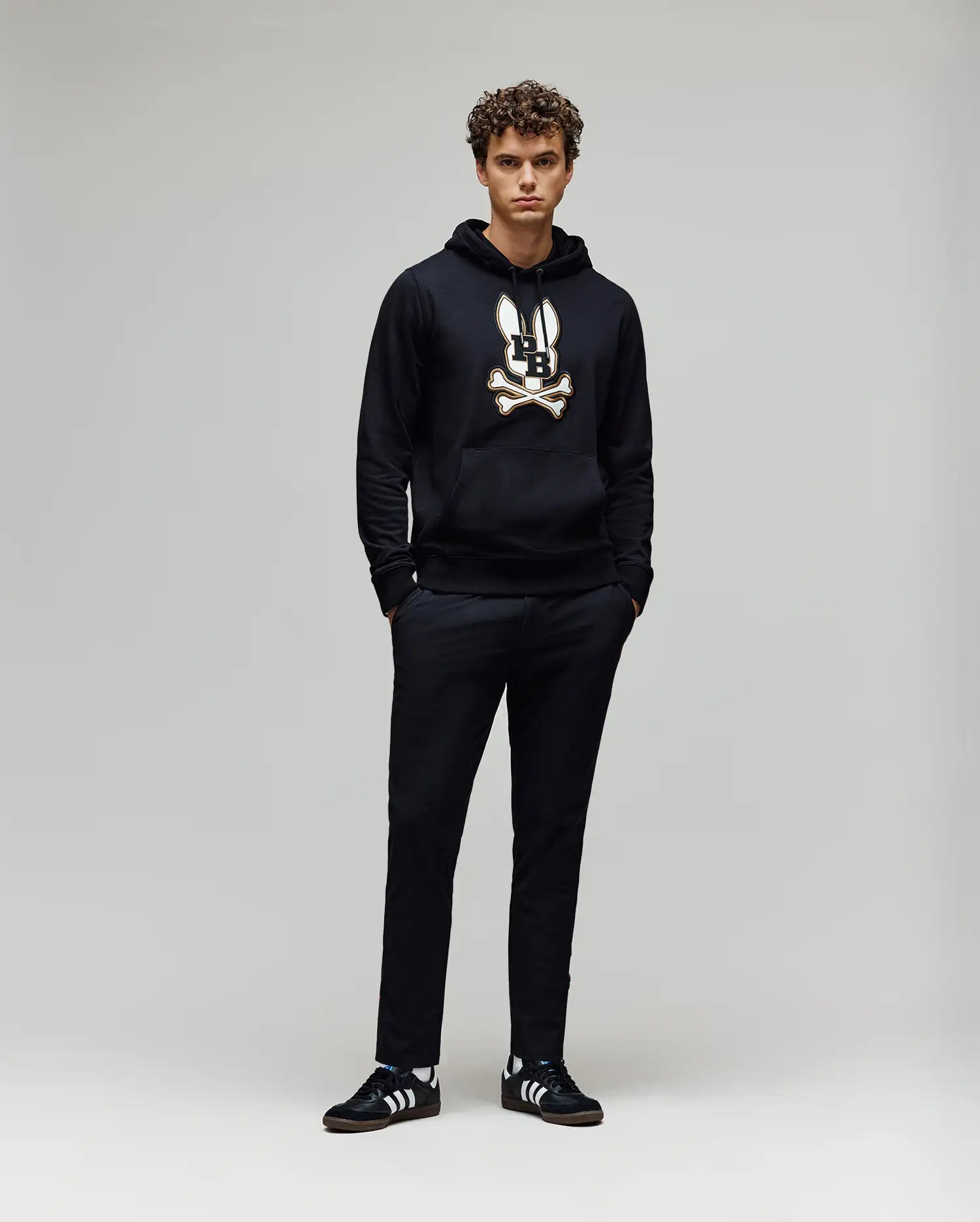 A young man with curly hair stands against a plain light background, hands in his pockets, wearing the Psycho Bunny MENS WADE HOODIE (MT0500112) featuring a skull and crossbones graphic, paired with black pants and black Adidas sneakers with white stripes.