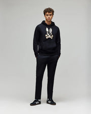 A young man with curly hair stands against a plain light background, hands in his pockets, wearing the Psycho Bunny MENS WADE HOODIE (MT0500112) featuring a skull and crossbones graphic, paired with black pants and black Adidas sneakers with white stripes.