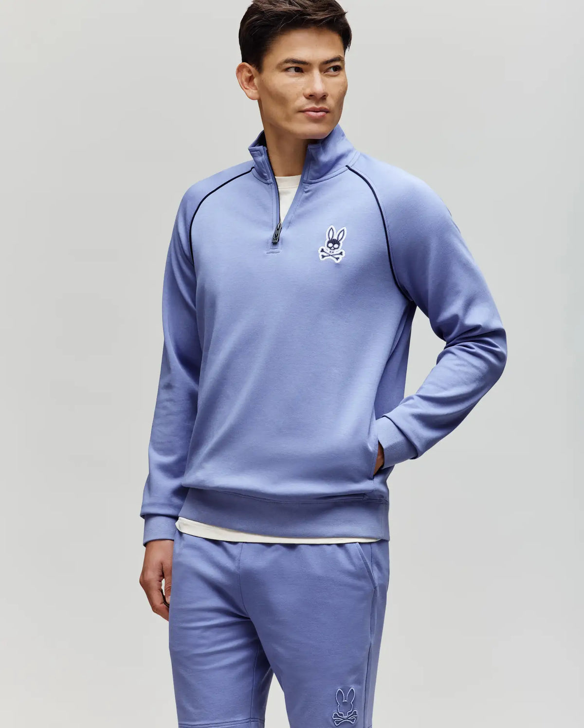 A man stands against a light gray background wearing the Psycho Bunny MENS SHAWN QUARTER ZIP SWEATSHIRT (MT0500115) in light purple with matching white bunny logo shorts, looking to the side with his hands in his pockets.