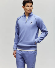 A man stands against a light gray background wearing the Psycho Bunny MENS SHAWN QUARTER ZIP SWEATSHIRT (MT0500115) in light purple with matching white bunny logo shorts, looking to the side with his hands in his pockets.