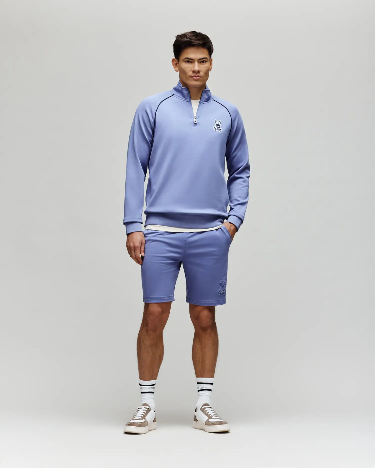 A man wears the Psycho Bunny MENS SHAWN QUARTER ZIP SWEATSHIRT (MT0500115) in light purple, featuring a 1/4 zip and embroidered logo, with matching shorts, white socks with black stripes, and white sneakers against a plain background.