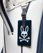 A close-up of the Psycho Bunny OUTLINE BUNNY GOLF TOUR BAG - PBMG4FC1 in white and dark blue, featuring a leather strap and a dark tag with a rabbit-eared skull and crossbones symbol.