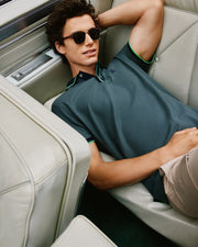 A person sporting the dark, stylish Psycho Bunny MENS KENT PIQUE POLO SHIRT - B6K951D200 and sunglasses reclines comfortably on cream-colored car seats, with one arm resting behind their head. The scene exudes a relaxed and fashionable vibe.