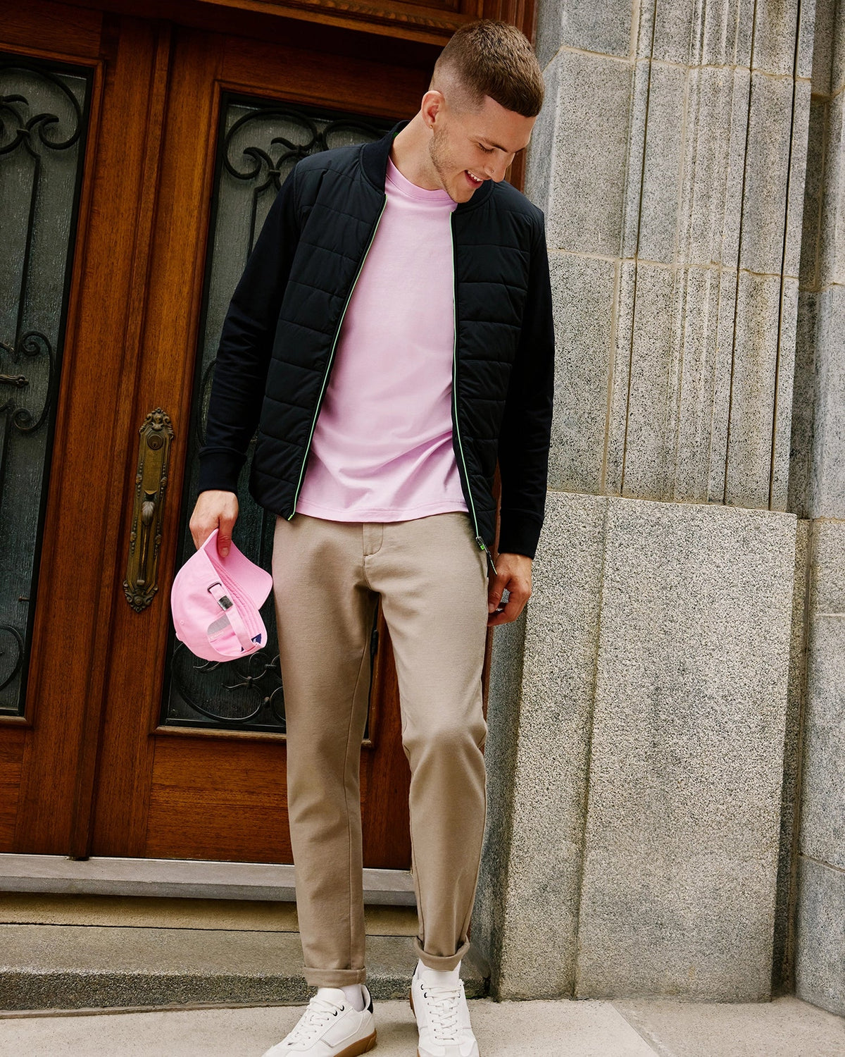 A person stands in front of a wooden door, wearing the Psycho Bunny MENS MYKEL MIXED MEDIA JACKET in black over a pink shirt, complemented by beige pants and white sneakers. They hold a pink cap in one hand and gaze downwards amidst a building with stone walls and decorative glass accents.