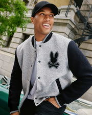 A person wearing a black cap and the stylish MENS TOM WOOL BOMBER JACKET - B6J744D200 from Psycho Bunny, adorned with a bunny patch, smiles while sitting on a car. In the background, there is a building with a fire escape surrounded by greenery.