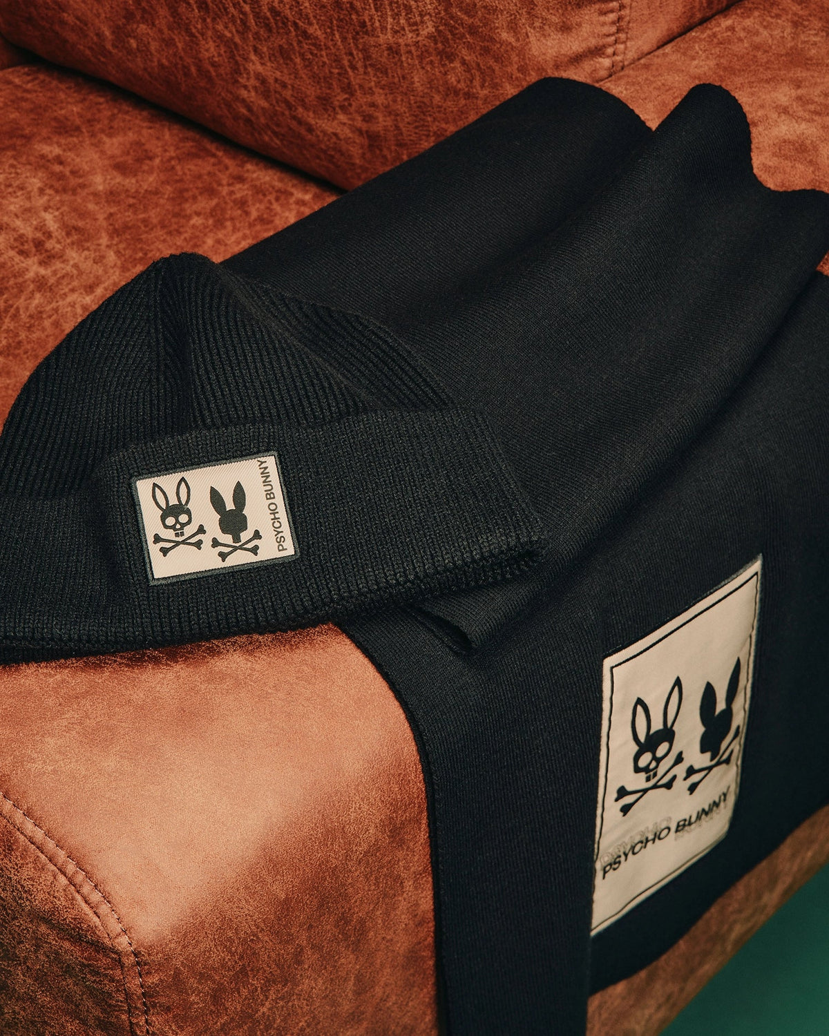 A luxurious accessory ensemble featuring the MENS MONROE TWILL PATCH SCARF from Psycho Bunny, crafted from a cotton wool blend, combined with a stylish black beanie adorned with an elegant twill patch showcasing the iconic bunny and crossbones design, rests elegantly on a brown leather chair.