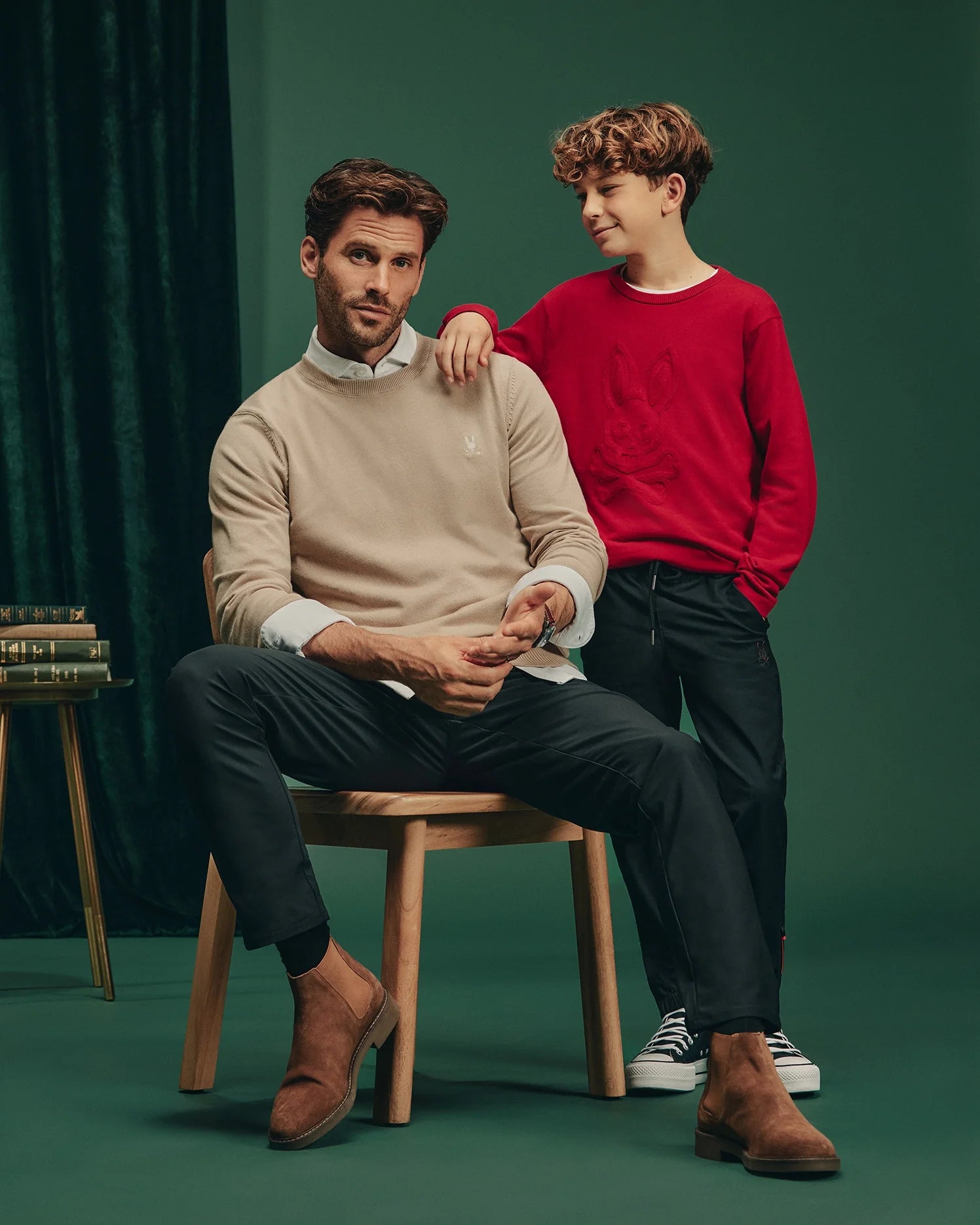 A man in a beige sweater sits on a stool, gazing ahead, while a boy in a red KIDS COORY CHAINSTITCH COTTON SWEATER by Psycho Bunny stands next to him with his hand on the man's shoulder. Both wear dark pants and shoes against a dark green background.