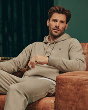 A person with wavy, short hair is sitting on a brown leather chair, wearing a beige hoodie and the Psycho Bunny MENS LEVY ESSENTIAL FLEECE SWEATPANT - B6P721D200. The background features a dark curtain. The person has a thoughtful expression.