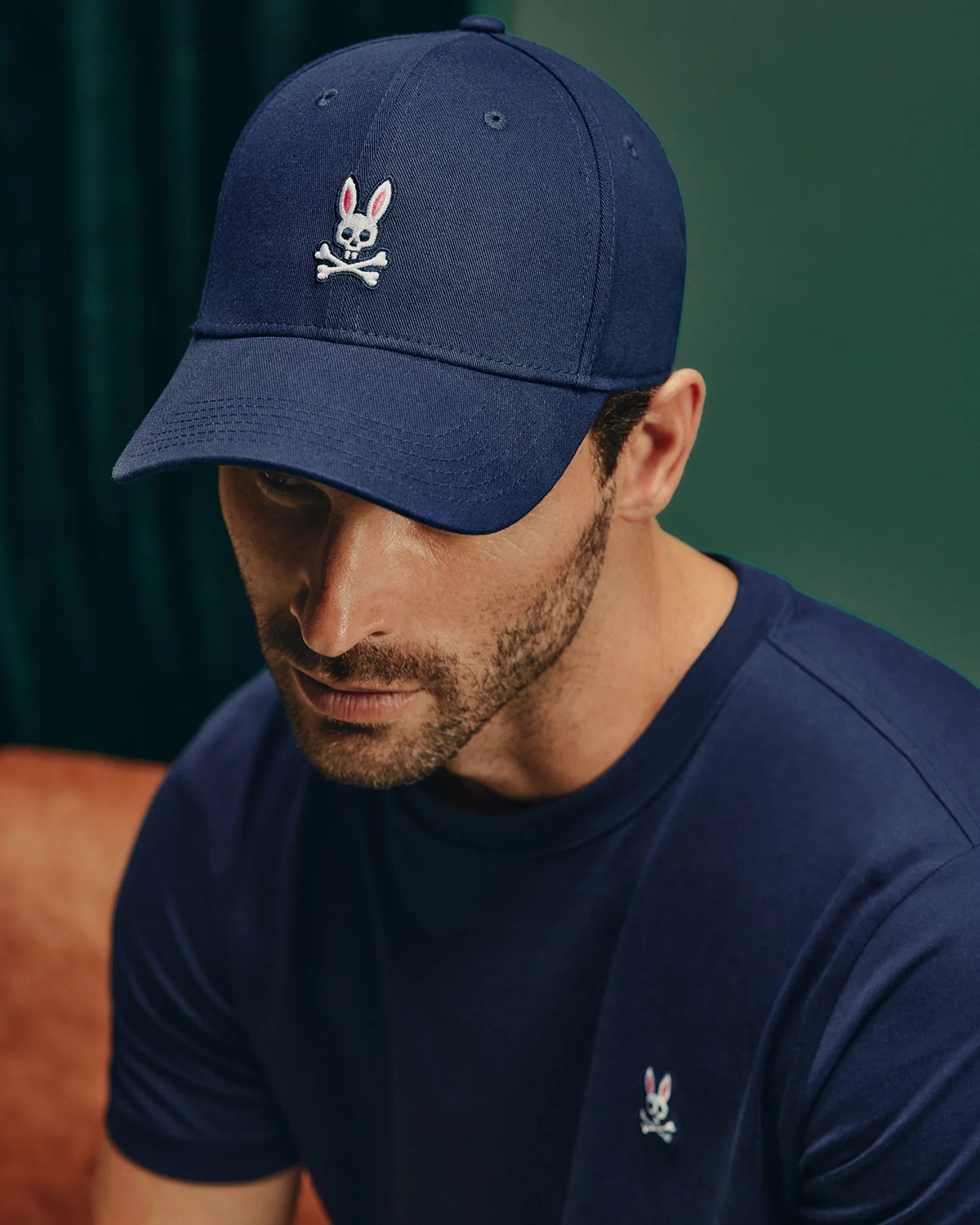 A man wears the Psycho Bunny MENS CLASSIC BASEBALL CAP - B6A816A1HT in navy blue, featuring a skull and crossbones logo with bunny ears, made from cotton twill for an adjustable fit. He pairs it with a matching navy blue t-shirt adorned with a small Psycho Bunny logo. The scene is set against a dark green curtain and brown leather surface.