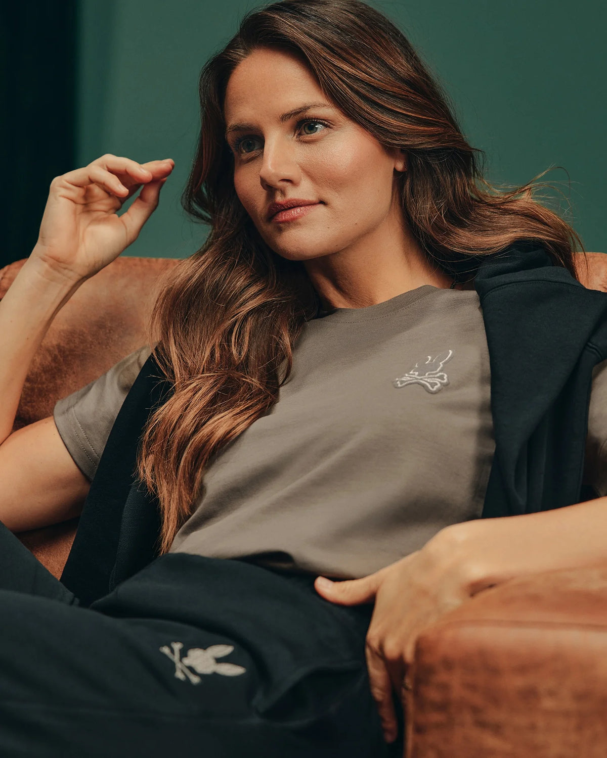 A person with long brown hair sits thoughtfully on a brown leather couch, wearing the Women's Alexander Graphic Tee by Psycho Bunny in grey, featuring a small logo. They rest their left arm on the armrest and hold their right hand near their face, with a slight smile. The background is green.