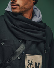 The individual wears a dark coat adorned with an elegant Psycho Bunny patch featuring two stylized bunny faces. To complement the coat, their Monroe Twill Patch Scarf by Psycho Bunny showcases a charming bunny design. Completing the ensemble are a hoodie and black beanie, all set against a vibrant green background.