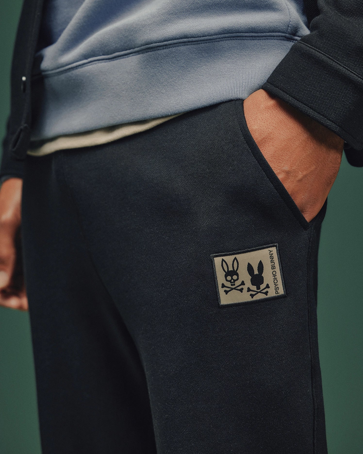 The individual stands with one hand in the pocket of Psycho Bunny's MENS MONROE TWILL PATCH FLEECE SWEATPANT, featuring a distinctive bunny skull and crossbones patch. Their light blue hoodie enhances the ensemble against a dark green backdrop.