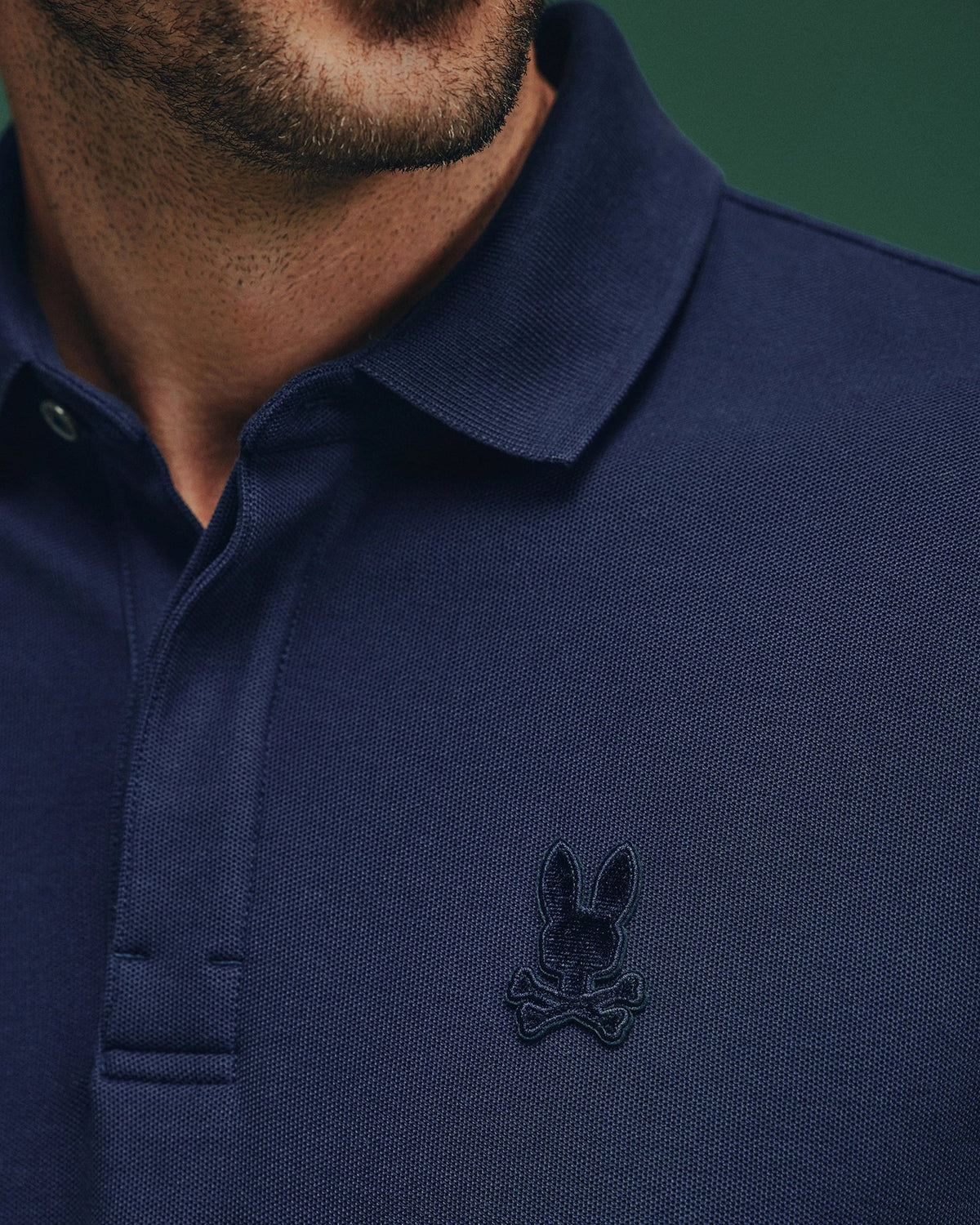 Close-up of a person wearing the MENS BRANSON PIQUE POLO - B6K108E200 by Psycho Bunny, featuring a navy blue design with a Velvet Patch Logo showcasing a bunny and crossbones emblem on the chest. The background is dark green.