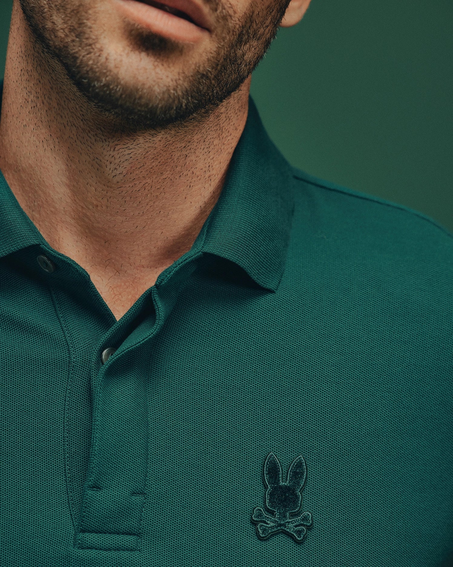 A man wearing the MENS BRANSON PIQUE POLO - B6K108E200 by Psycho Bunny, in a premium green hue with a black embroidered bunny and crossbones logo on the chest, stands against a matching green background. The image emphasizes the upper torso and part of the face without fully revealing facial features, effectively showcasing this classic men's Pima cotton polo.