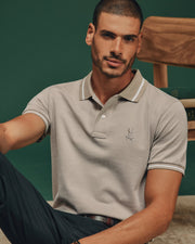 A man wearing a light gray Psycho Bunny MENS WARSAW JACQUARD PIQUE POLO - B6K901E200, featuring the brand's signature logo, casually leans against a wooden chair amidst a green backdrop. With short hair and a beard, he portrays an air of relaxed elegance.