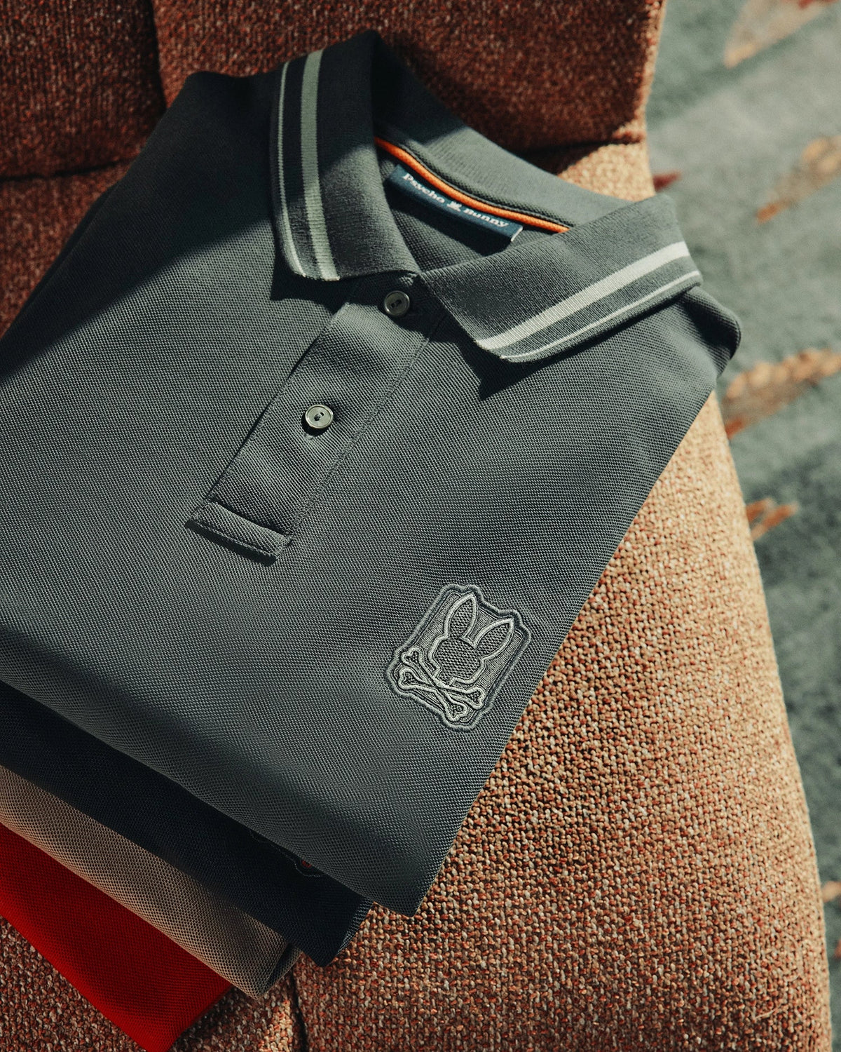 A meticulously folded MENS PIERCE PIQUE POLO from Psycho Bunny, crafted from luxurious Pima cotton, showcases an embroidered Bunny logo on the chest. This dark polo shirt features a white-striped collar and is arranged on a textured surface, gently illuminated by sunlight spotlighting its fabric.