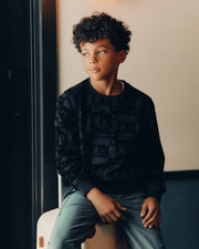 A young boy with curly hair sits on a light-colored bench, wearing the KIDS ROMEO LIGHTWEIGHT SWEATSHIRT by Psycho Bunny and light gray pants. He gazes thoughtfully to the side, surrounded by softly lit warm tones.