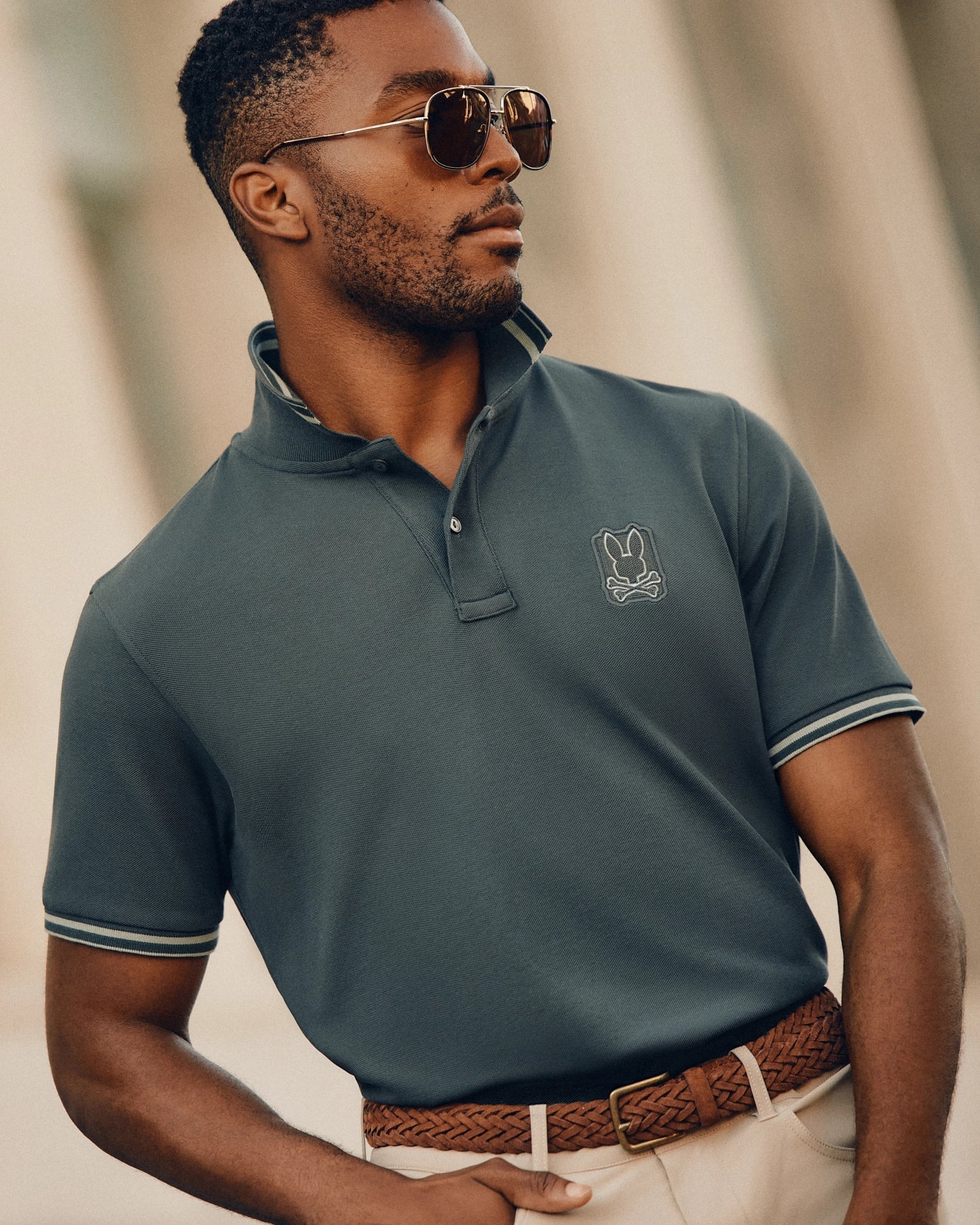 A man wearing the Psycho Bunny MENS PIERCE PIQUE POLO - B6K201E200, complete with an embroidered Bunny logo, stands confidently outdoors in sunglasses. His dark Pima cotton polo shirt is neatly tucked into beige pants, cinched by a braided brown belt. The softly blurred background highlights the man's stylish appearance.