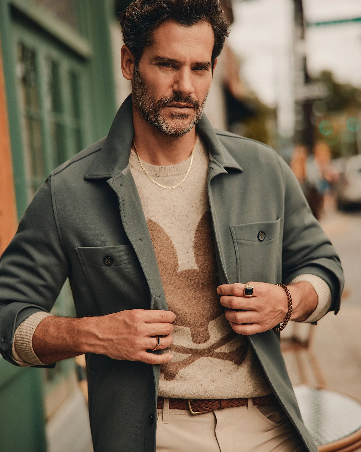A bearded man stands confidently on a street, exuding an urban vibe. He wears the stylish Psycho Bunny Men's Bedford Shirt Jacket - B6J290B200 over a cotton-blend sweater with a rabbit design, paired with beige pants and accessories including a necklace, rings, and a bracelet. The backdrop features a green door and blurred city buildings.