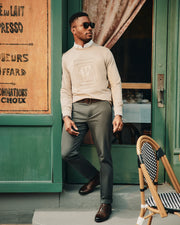 A stylish man wearing sunglasses leans against a green storefront, donning the Psycho Bunny MENS OAK CITY CREW NECK - B6S274E200 in beige. The ensemble is completed with a white shirt, gray pants, and brown shoes. Nearby, a wicker chair sits by the window partially veiled by a pink curtain.