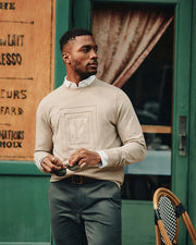 Wearing the premium MENS OAK CITY CREW NECK sweater by Psycho Bunny, a man stands confidently in front of a green door and sign, holding sunglasses. A bistro chair and curtain add charm to the scene, completing the elegant snapshot.