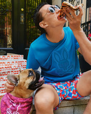 A person wearing sunglasses and a blue t-shirt adorned with the vibrant Psycho Bunny logo sits on a stoop, munching on a slice of pizza while holding a box. A French Bulldog sporting a pink bandana looks up at them. In the background, two more pizza boxes are stacked neatly beside them.
