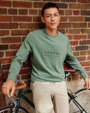 A man wearing the MENS TACOMA SWEATSHIRT - B6S927D200 by Psycho Bunny, in green, sits on a bicycle exuding luxurious comfort. He is posed against a brick wall, sporting beige pants with premium stretch and smiling at the camera.