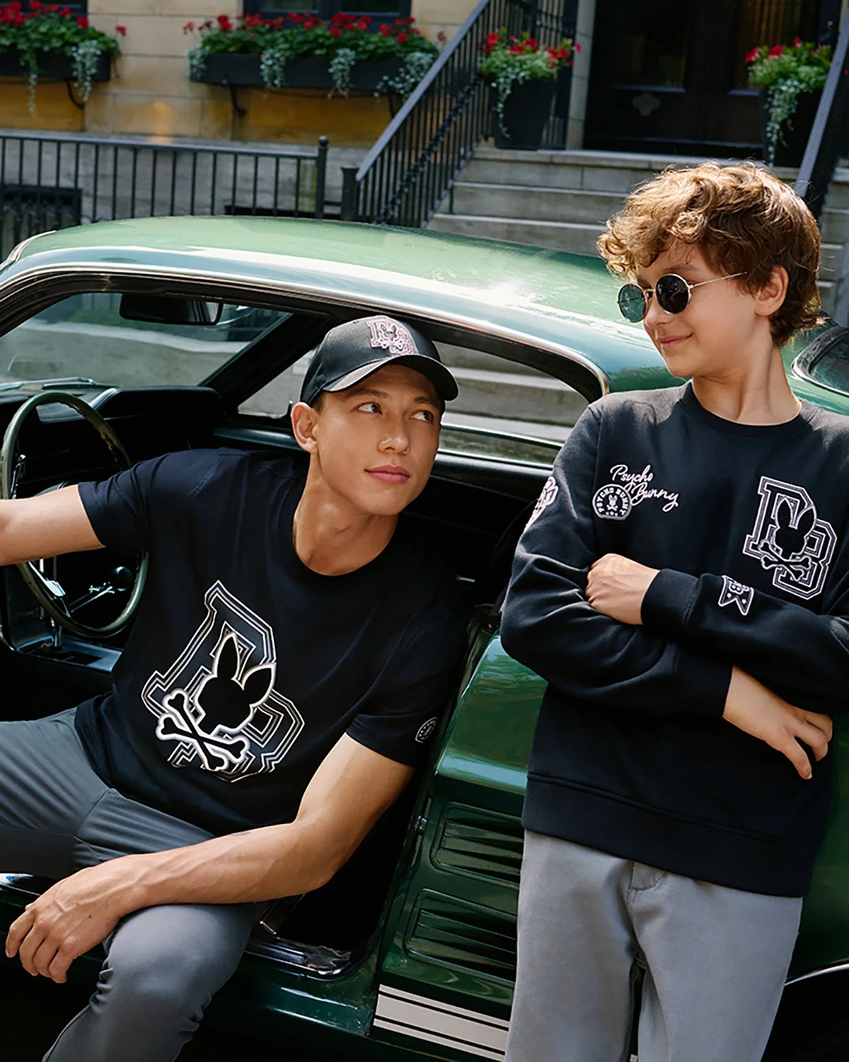 There are two individuals by a green car. The one on the left is seated inside, donning a black cap and t-shirt. The one on the right stands outside, smiling while wearing sunglasses and the Psycho Bunny KIDS ZACK VARSITY SWEATSHIRT with chenille embroidery. Both are in casual outfits featuring matching logos.