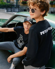 Two young men are sitting casually near a green car. One is seated inside, wearing a black cap and T-shirt, looking at the camera, while the other stands nearby wearing round sunglasses and a Psycho Bunny KIDS ZACK VARSITY SWEATSHIRT - B0S880D200 with chenille embroidery on the back.