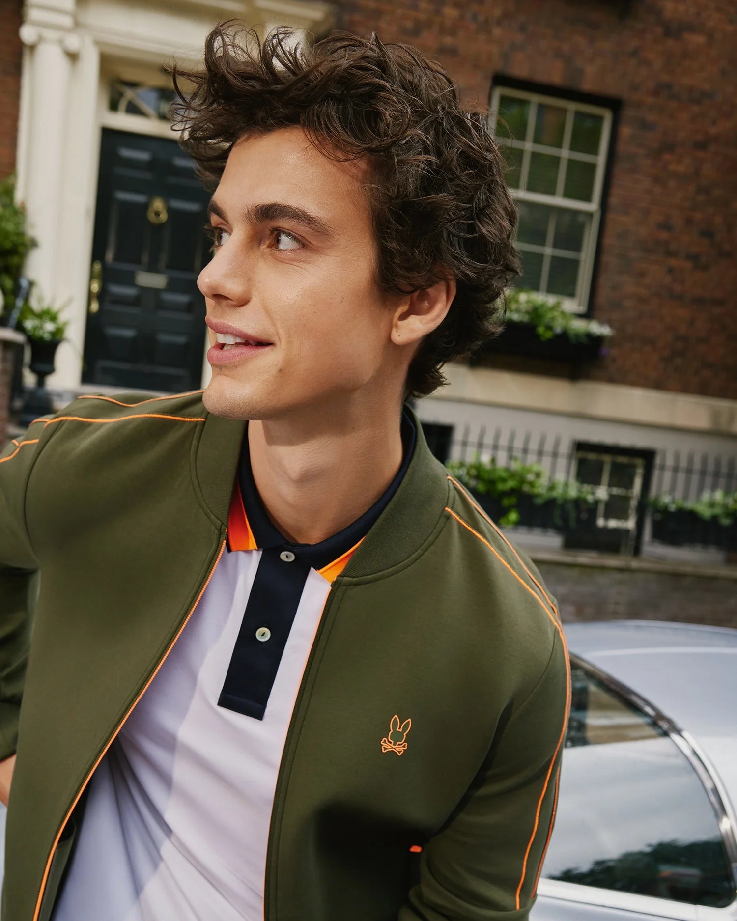 A young man with curly hair looks to the side and smiles. He sports the high-quality MENS TRAVIS TRACK JACKET - B6J142D200 by Psycho Bunny, featuring orange trim, over a white and navy polo shirt. Behind him is a car and a brick building with a dark door and potted plants on the windowsills.