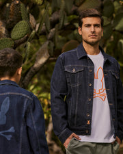 Two men wearing the MENS PATHE DENIM JACKET - B6J741X1CO by Psycho Bunny are standing outdoors near a cactus. The man facing the camera sports a white shirt with an orange logo, while the other, highlighting embroidered details on his jacket, has his back to the camera. Trees and greenery can be seen in the background.