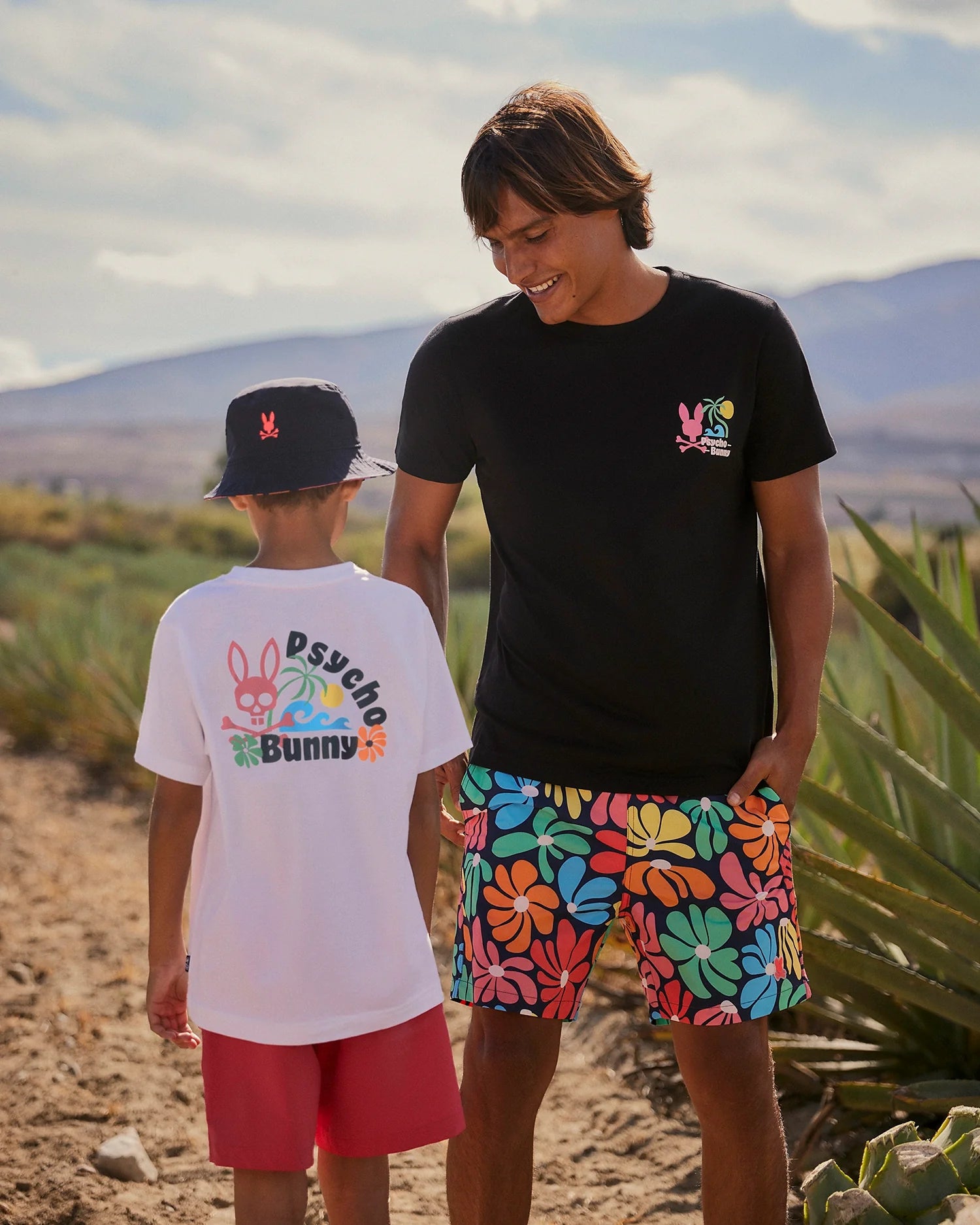 Two people stand outdoors on a sunny day, both wearing bright Psycho Bunny clothing. One, an adult in a black shirt and colorful MENS MENTZ SWIM TRUNK - B6W320B2SW by Psycho Bunny, faces and smiles at the other, a child in a white T-shirt and red shorts. There are plants and mountains in the background.