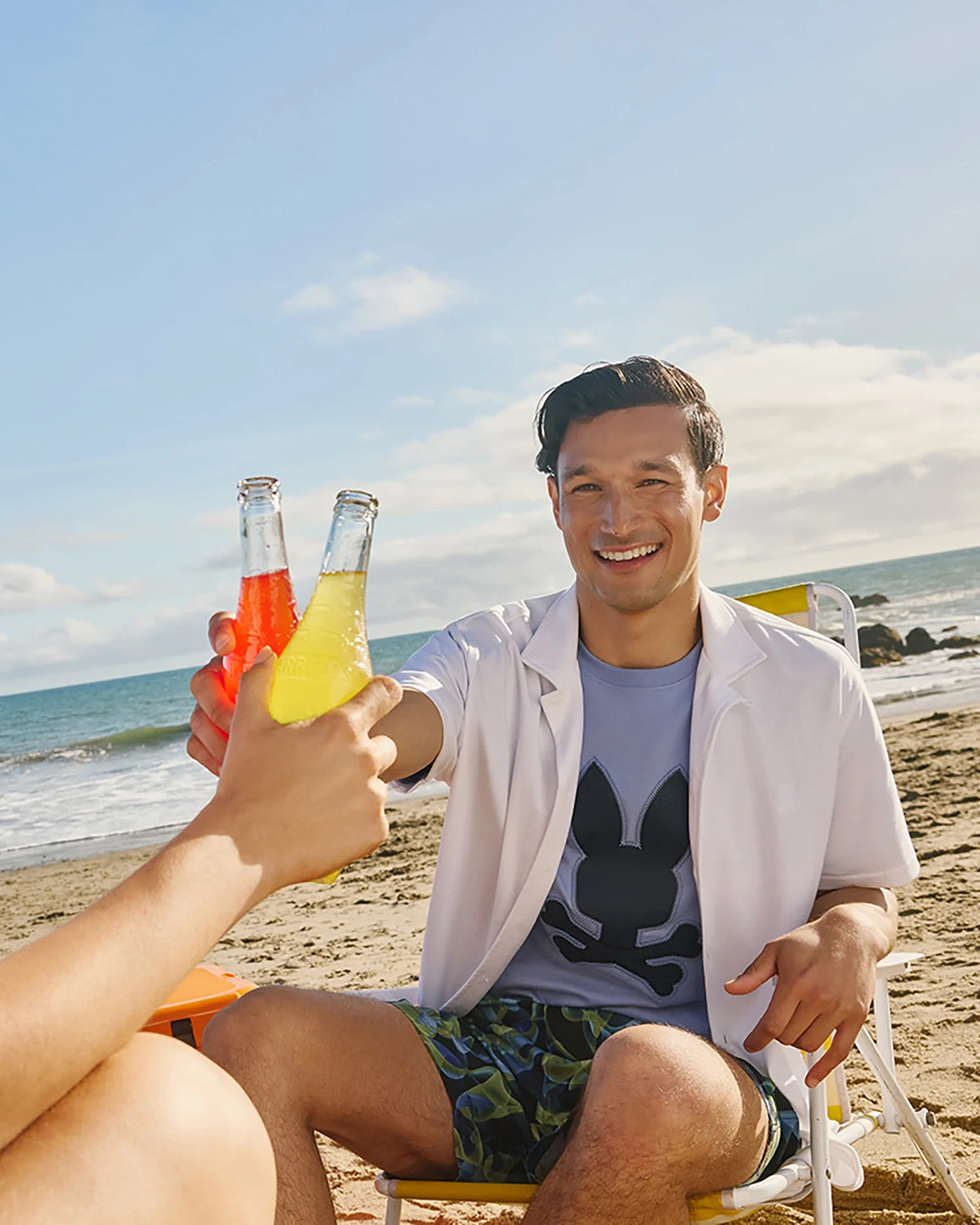 Two people toast with bottles of colorful drinks as they unwind on a sandy beach. One person, lounging in a beach chair and wearing Psycho Bunny's MENS GOLDCREEK ALL OVER PRINT SWIM TRUNK - B6W672C200, sports a white shirt over a graphic tee and patterned shorts. The scene is set against the backdrop of the ocean under a partly cloudy sky, evoking a sunny and cheerful day.