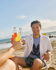 Two people toast with bottles of colorful drinks as they unwind on a sandy beach. One person, lounging in a beach chair and wearing Psycho Bunny's MENS GOLDCREEK ALL OVER PRINT SWIM TRUNK - B6W672C200, sports a white shirt over a graphic tee and patterned shorts. The scene is set against the backdrop of the ocean under a partly cloudy sky, evoking a sunny and cheerful day.